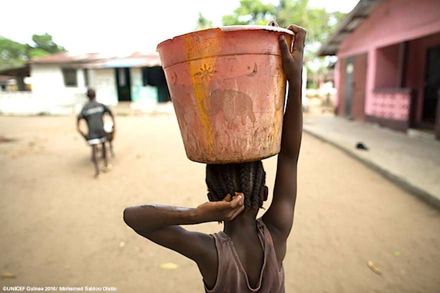 Haciendo lo que muchas niñas aún tienen que hacer en Guinea, Princess Johnson, de 8 años, lleva a casa un gran recipiente con agua para su familia.